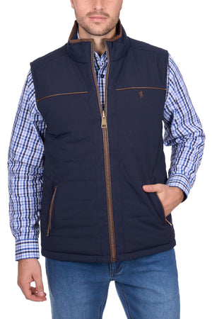 A man stands against a white background wearing the Thomas Cook Toby Revs Vest in blue with brown accents, over a blue checkered shirt and blue jeans, with one hand in his vest pocket.