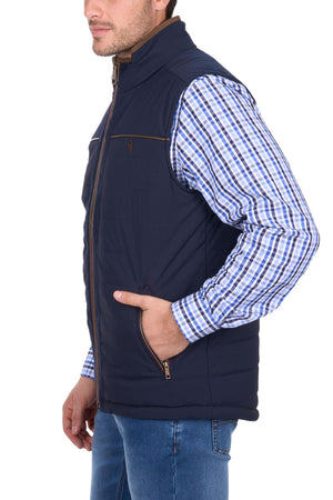 A man wears the Thomas Cook Toby Revs Vest, a navy blue quilted vest with zippered pockets, over a blue checkered shirt, standing with one hand in his pocket against a white background.