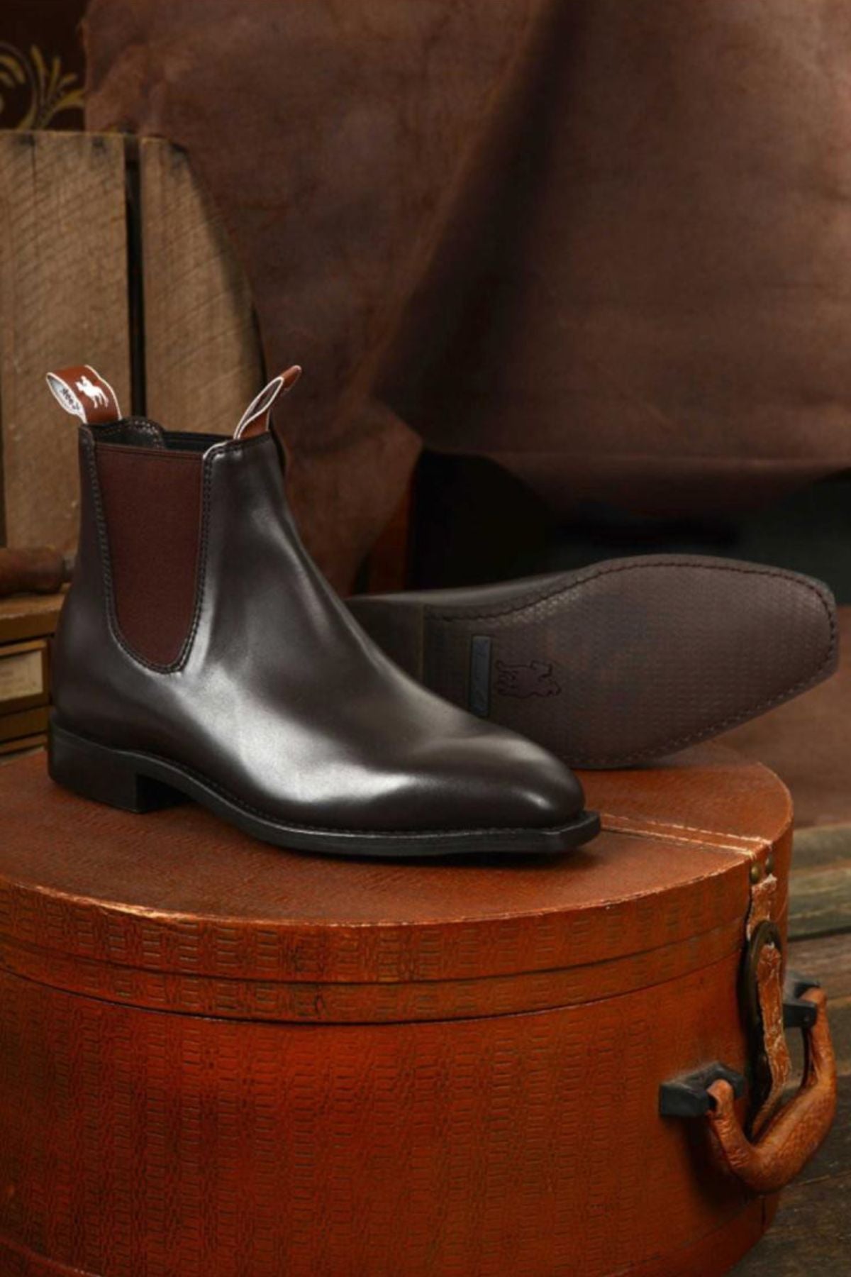 The Thomas Cook Trentham Cushion Tech Dress Boot, a pair of black premium leather Chelsea boots with brown elastic side panels, is shown on a round textured brown case with wooden crates behind. Made in Australia.