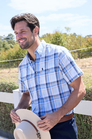 A smiling man stands outdoors in a Thomas Cook Brian Short Sleeve Shirt with blue and white plaid, holding a light-colored wide-brimmed hat. Greenery and a white fence are visible in the background.