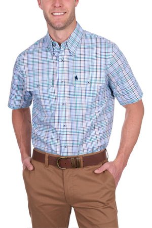 A man wears the Thomas Cook Matt Short Sleeve Shirt—a button-up cotton shirt with blue, green, and white plaid—tucked into tan pants with a brown belt. Smiling, he has his hands in his pockets.