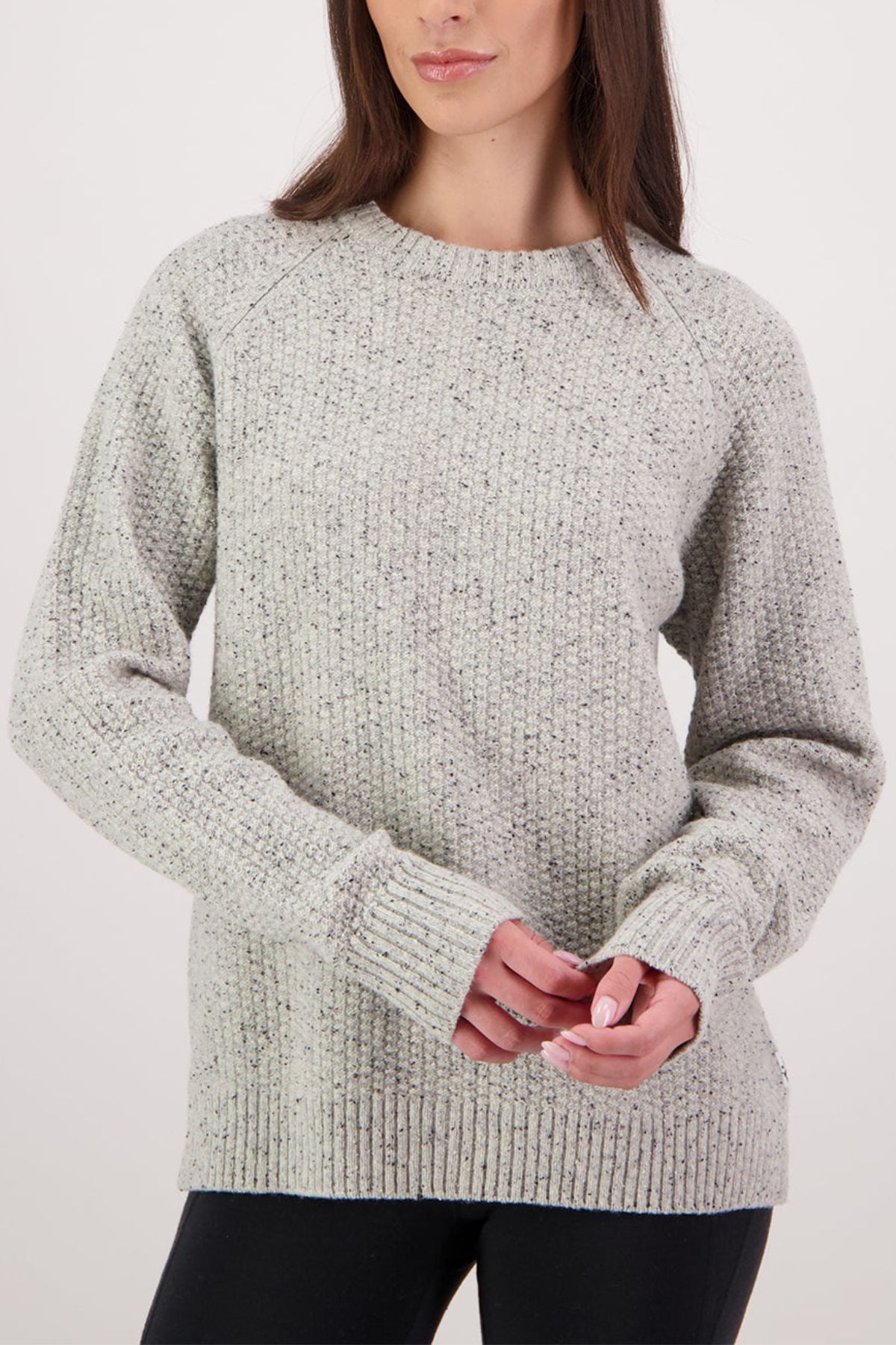 A woman stands against a plain white background, facing forward with hands loosely clasped, wearing the Swanndri Lyttelton Womens Waffle Crew—a light gray ribbed knit sweater with long sleeves—paired with black pants for a cozy look.