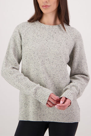A woman stands against a plain white background, facing forward with hands loosely clasped, wearing the Swanndri Lyttelton Womens Waffle Crew—a light gray ribbed knit sweater with long sleeves—paired with black pants for a cozy look.