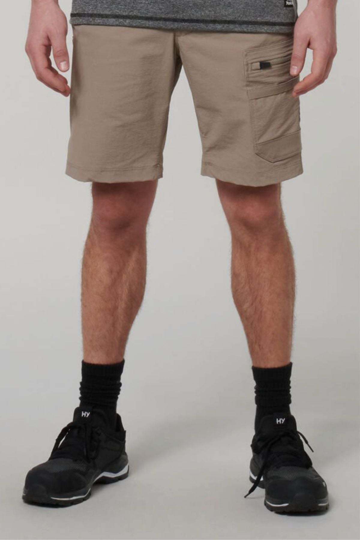 A person stands against a plain light background, wearing Hard Yakka X Range Mid Shorts in beige Cordura nylon, paired with black socks, black sneakers, and a partially visible gray shirt.