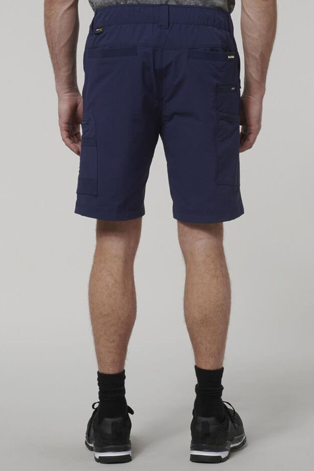 A person faces away from the camera wearing Hard Yakka X Range Mid Short in navy blue with utility pockets, paired with black socks and athletic shoes, standing against a plain light gray background.