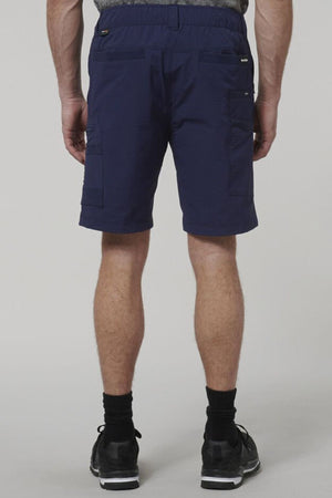 A person faces away from the camera wearing Hard Yakka X Range Mid Short in navy blue with utility pockets, paired with black socks and athletic shoes, standing against a plain light gray background.