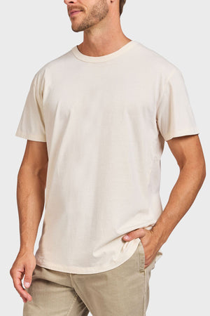 A man stands against a plain background with one hand in his pocket, wearing the Academy Brand Jimmy Tee in off-white and light beige pants. His face is partially out of frame.