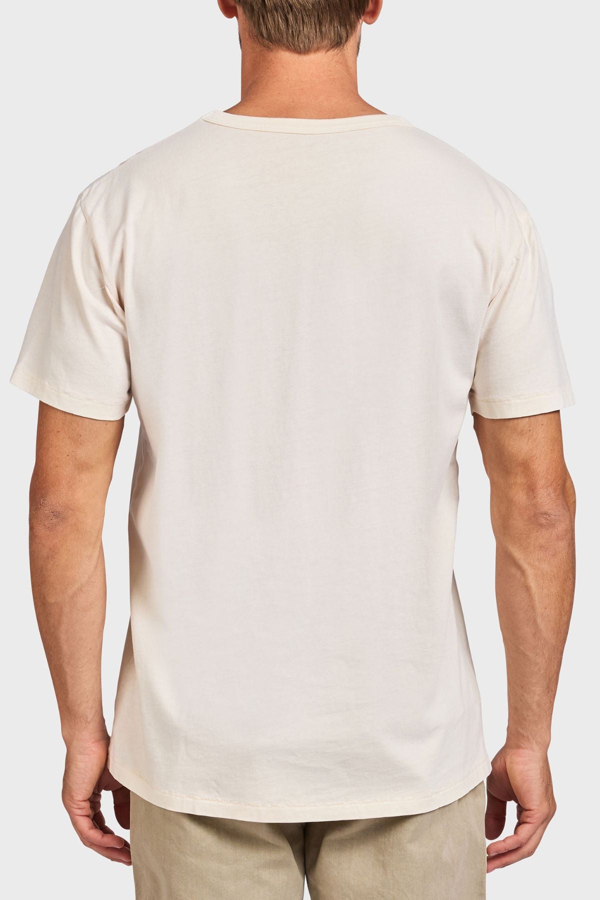 A man is seen from behind wearing the Academy Brand Jimmy Tee, a plain short-sleeved 100% cotton shirt, paired with light beige pants against a light gray background.