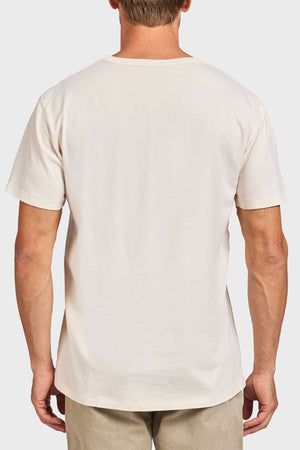 A man is seen from behind wearing the Academy Brand Jimmy Tee, a plain short-sleeved 100% cotton shirt, paired with light beige pants against a light gray background.