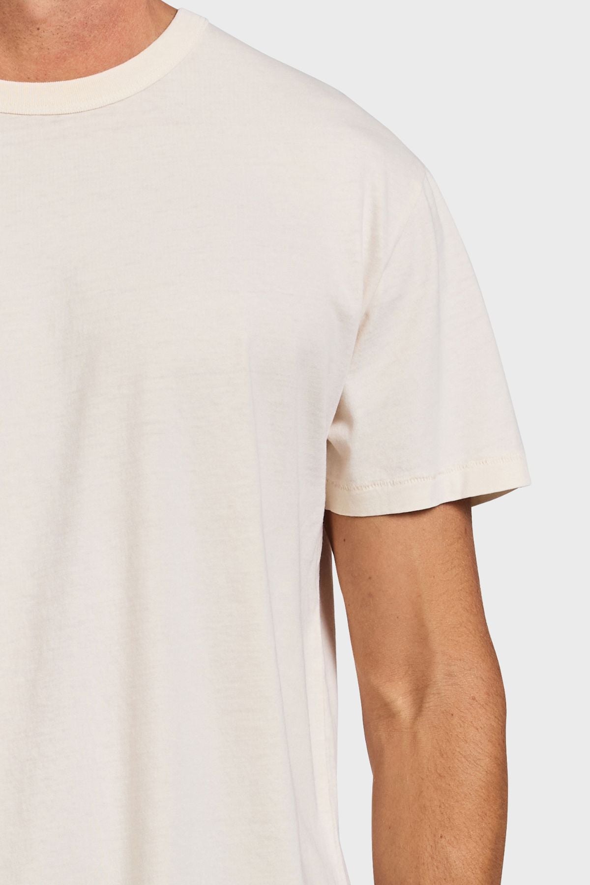 A close-up shows someone wearing the Academy Brand Jimmy Tee, a plain, short-sleeved, off-white 100% cotton top. Only the upper torso and part of the left arm are visible against a light gray background, highlighting its relaxed fit.