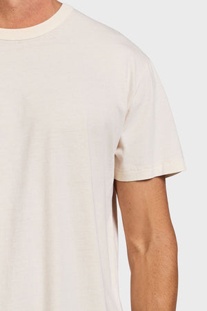 A close-up shows someone wearing the Academy Brand Jimmy Tee, a plain, short-sleeved, off-white 100% cotton top. Only the upper torso and part of the left arm are visible against a light gray background, highlighting its relaxed fit.