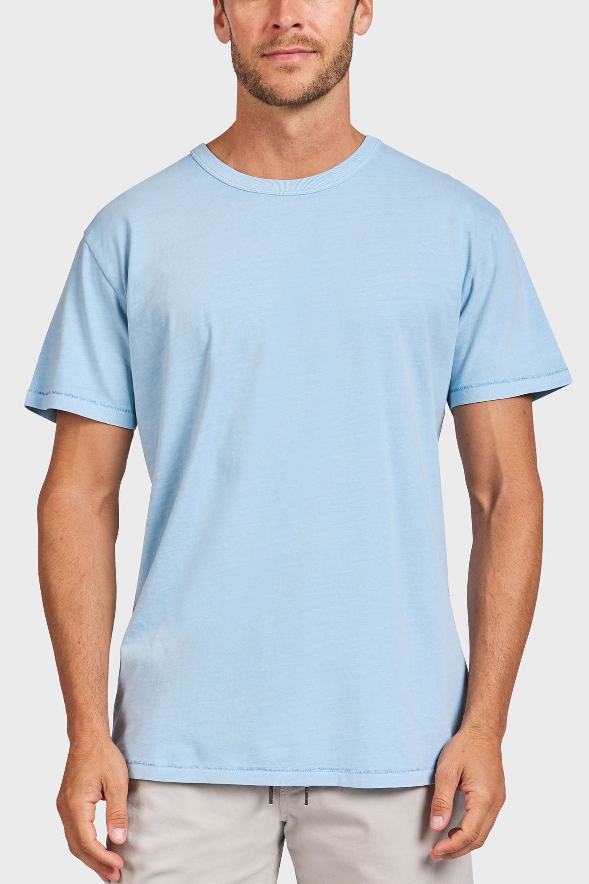 A man faces forward against a white background, wearing the Academy Brand Jimmy Tee in plain light blue and light-colored pants.