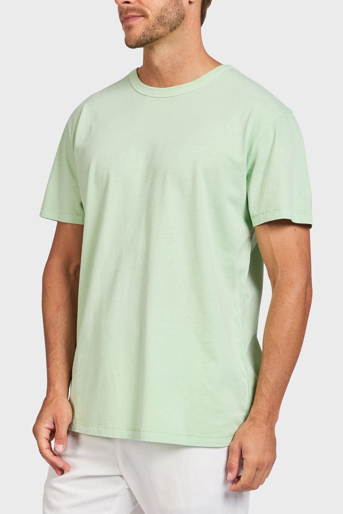 A man wears the Academy Brand Jimmy Tee in light green, a loose fit 100% cotton t-shirt, with white pants. He stands against a plain white background, his face partially turned out of the frame.