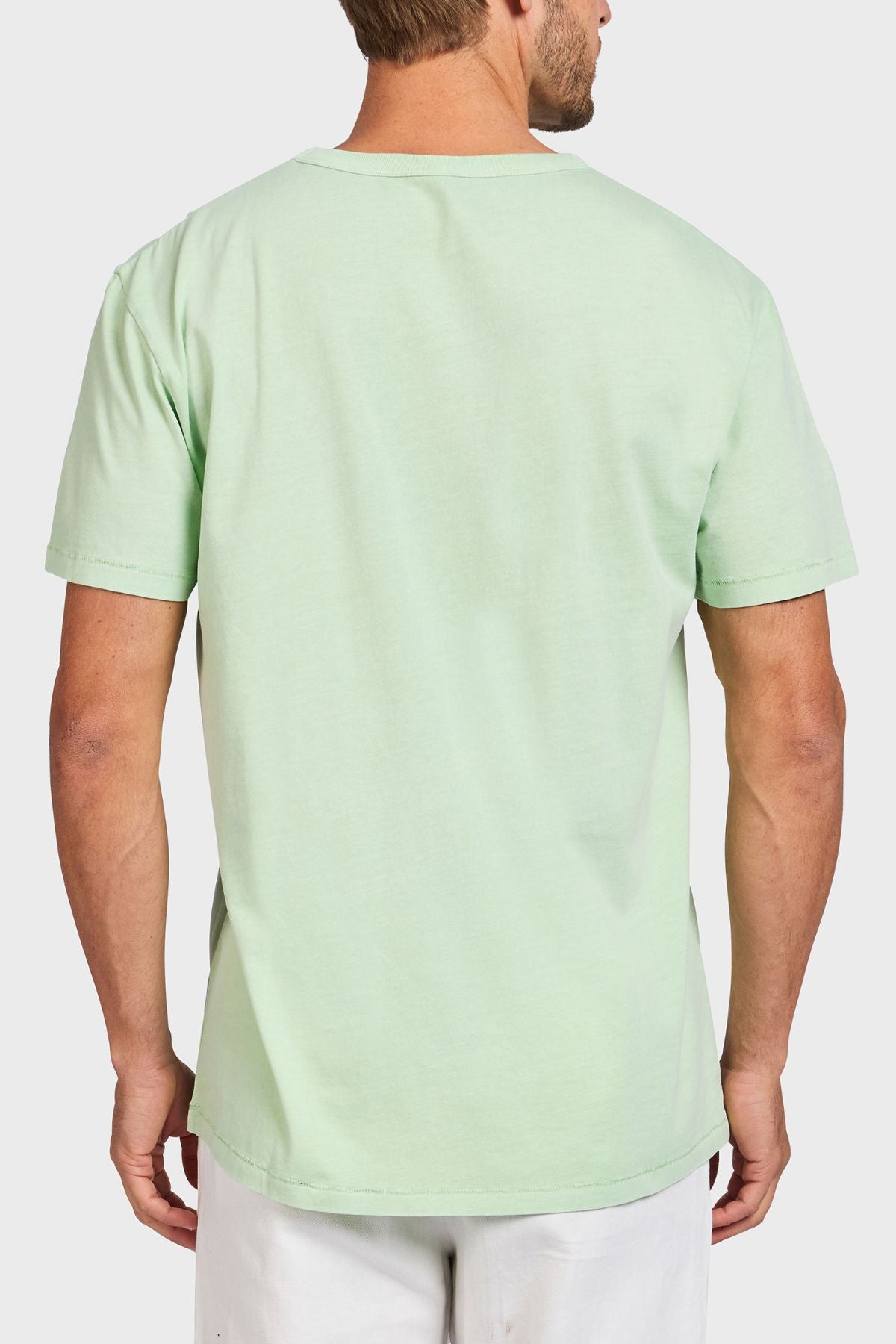A man is seen from behind wearing the Academy Brand Jimmy Tee, a loose-fit light green 100% cotton t-shirt, paired with white pants against a plain white background.