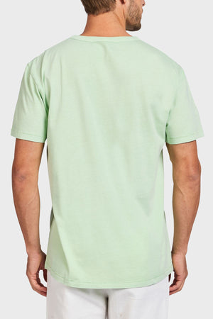A man is seen from behind wearing the Academy Brand Jimmy Tee, a loose-fit light green 100% cotton t-shirt, paired with white pants against a plain white background.