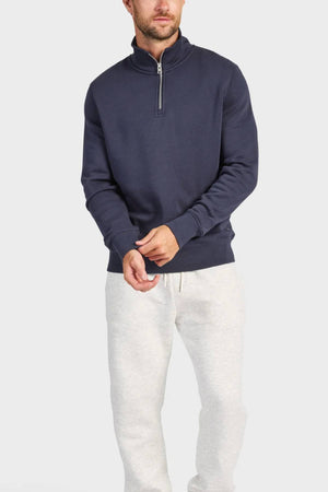 A man stands against a light background wearing the Academy Brand 1/4 Zip Sweat in navy blue with a vintage wash and light gray sweatpants. His head is slightly turned, and his hands are loosely clasped in front of him.