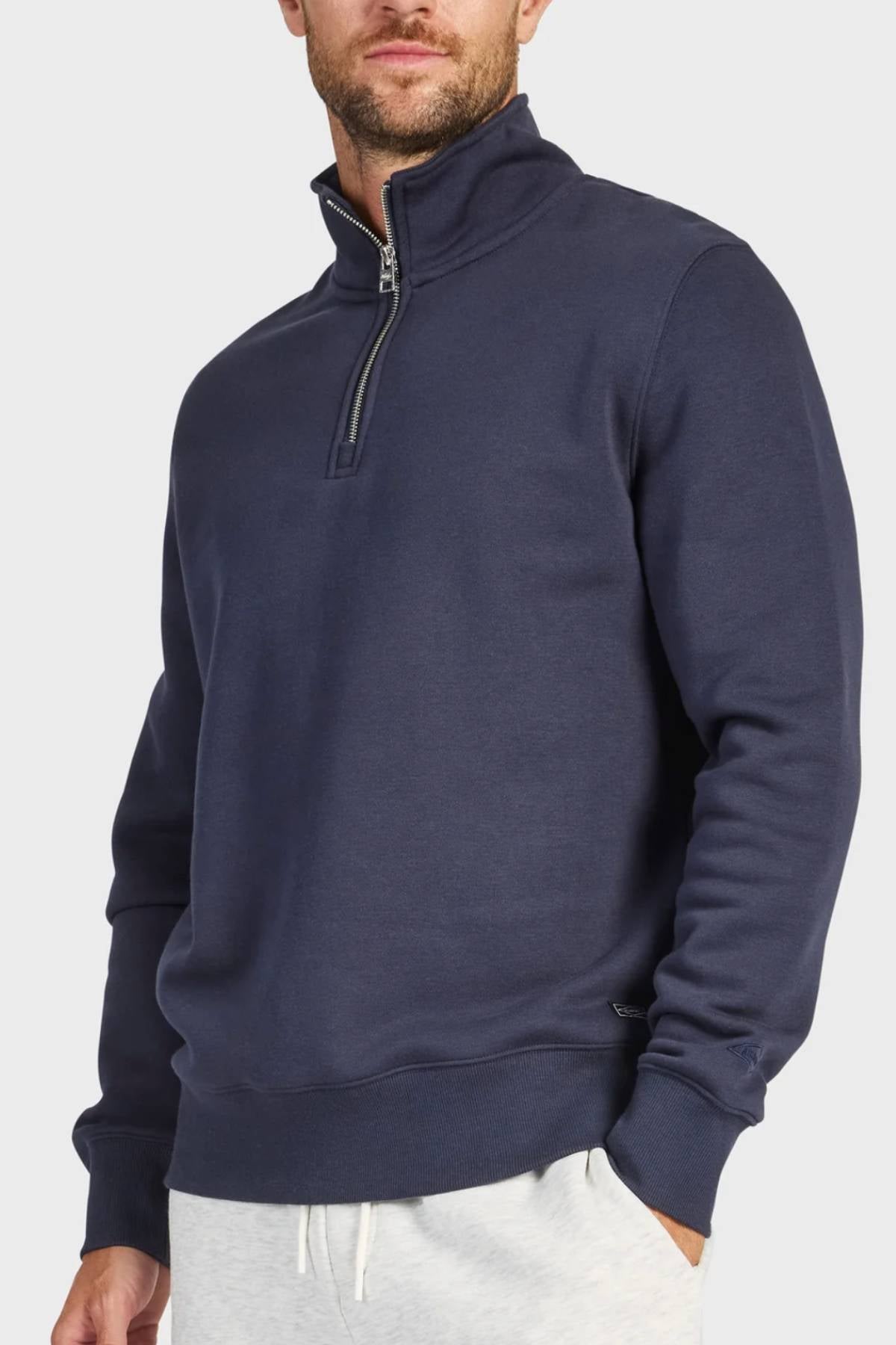 A man wears the Academy Brand 1/4 Zip Sweat in navy blue, featuring a vintage washed finish and stand-up collar, partially unzipped. He pairs it with light gray sweatpants against a plain light gray background. Made from premium brushed fleece.