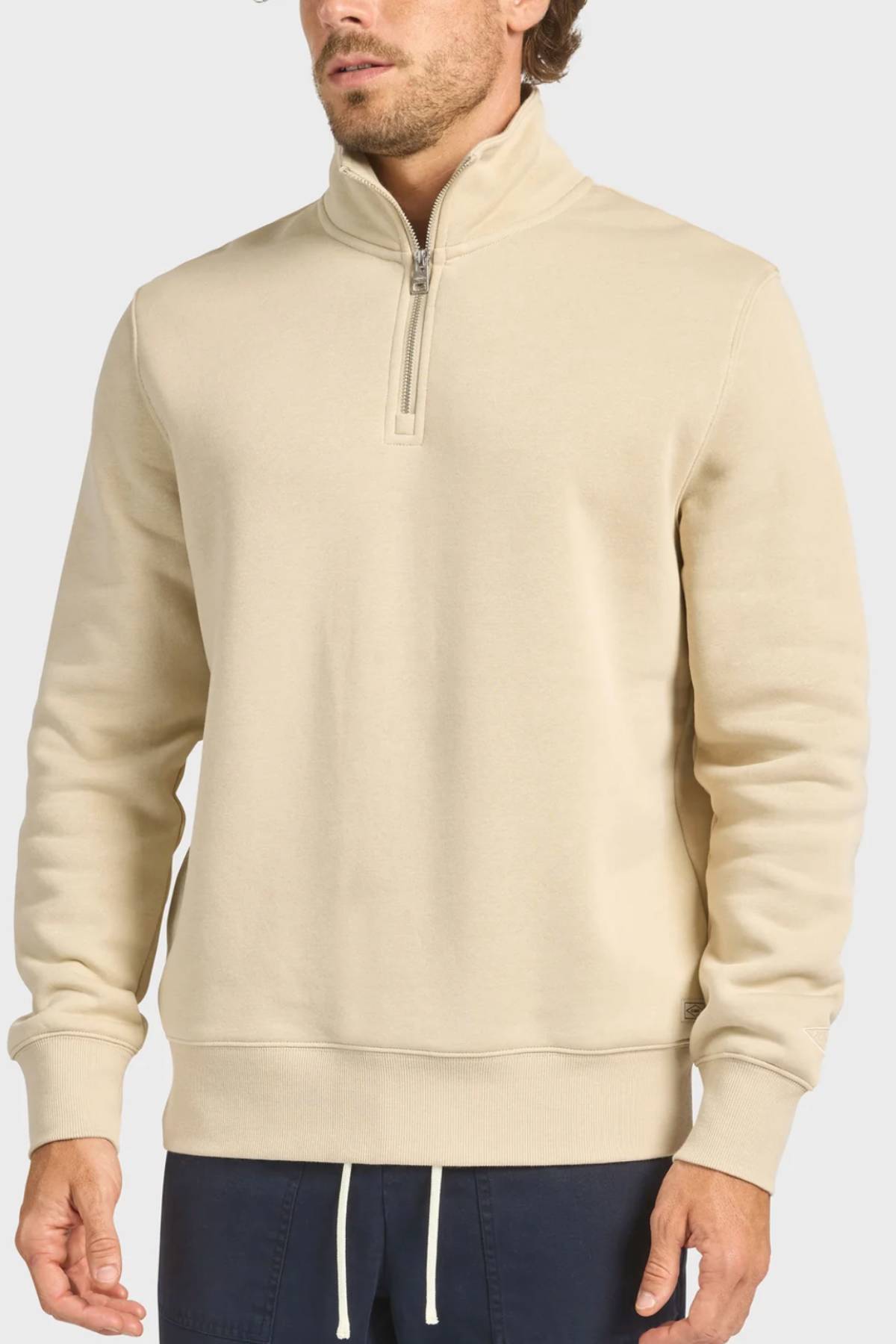 A man in the Academy Brand 1/4 Zip Sweat, featuring a vintage washed beige finish and ribbed cuffs, pairs it with dark blue drawstring pants while standing against a plain light background.