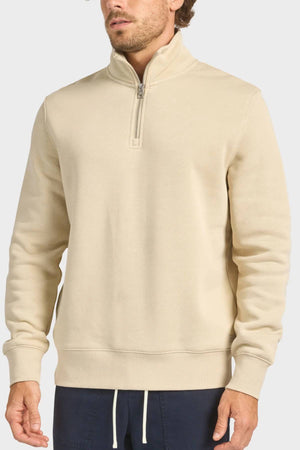 A man in the Academy Brand 1/4 Zip Sweat, featuring a vintage washed beige finish and ribbed cuffs, pairs it with dark blue drawstring pants while standing against a plain light background.