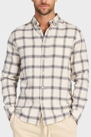 A man stands with his hands in the pockets of light-colored pants, wearing the Academy Brand Buhler Shirt—a cream shirt with a gray plaid pattern, yarn dye twill fabric, workwear buttons, a collar, and long sleeves.