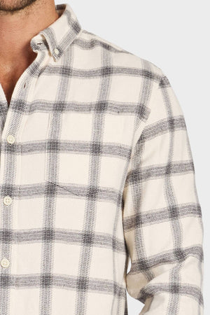 A person is shown from the upper torso up, wearing the Academy Brand Buhler Shirt—a cream button-up with black and gray plaid in yarn-dye twill. Only part of their left arm is visible.