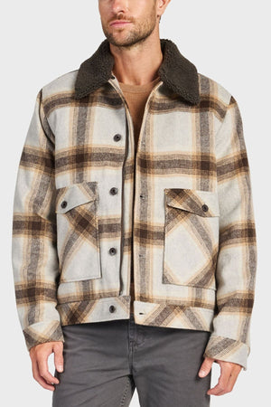 A man in the Academy Brand Conrad Jacket—a beige and brown plaid wool-blend jacket with a dark sherpa collar—layered over a tan sweater and paired with gray pants, stands before a plain light background.