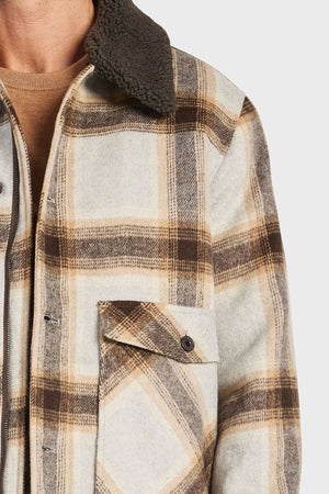 Close-up of a person wearing the Academy Brand Conrad Jacket—a brown, beige, and cream plaid wool blend with a soft dark collar and buttoned chest pocket. Only their neck, chest, and one arm are visible.