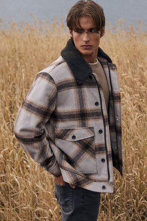 A young man stands in tall dry grass, wearing the Academy Brand Conrad Jacket—a lightweight plaid wool blend jacket featuring a dark collar—paired with a beige sweater and dark pants, looking to the side with a serious expression.