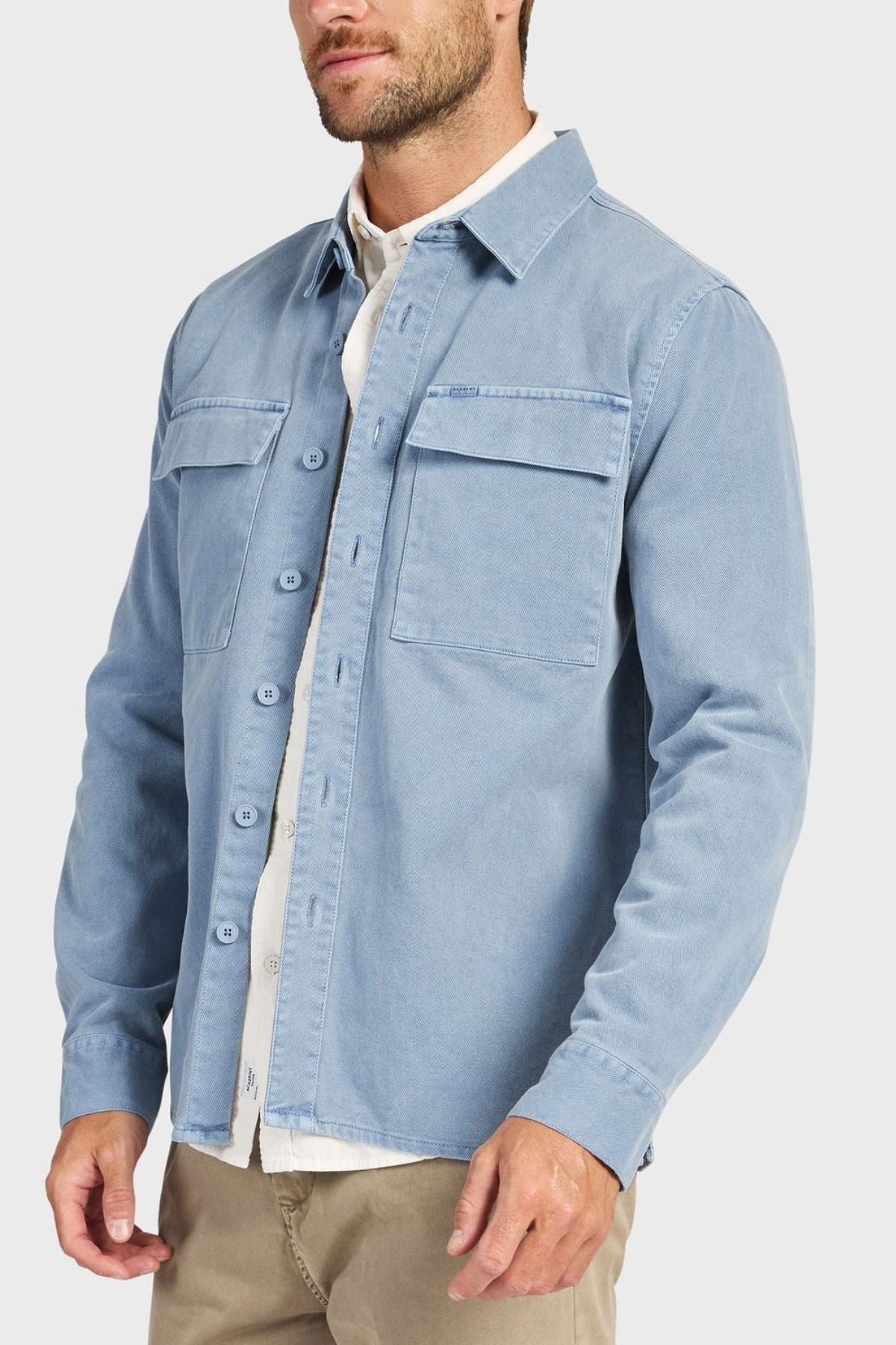A man wears the Academy Brand Essential Overshirt in light blue cotton twill with chest pockets, layered over a white shirt and beige pants, standing against a plain light background.
