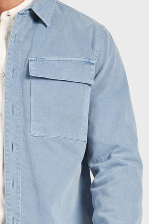 A close-up shows a person wearing the Academy Brand Essential Overshirt in light blue cotton twill with a flap chest pocket, layered over a white top. Only the left side of the shirt and part of the torso and arm are visible.