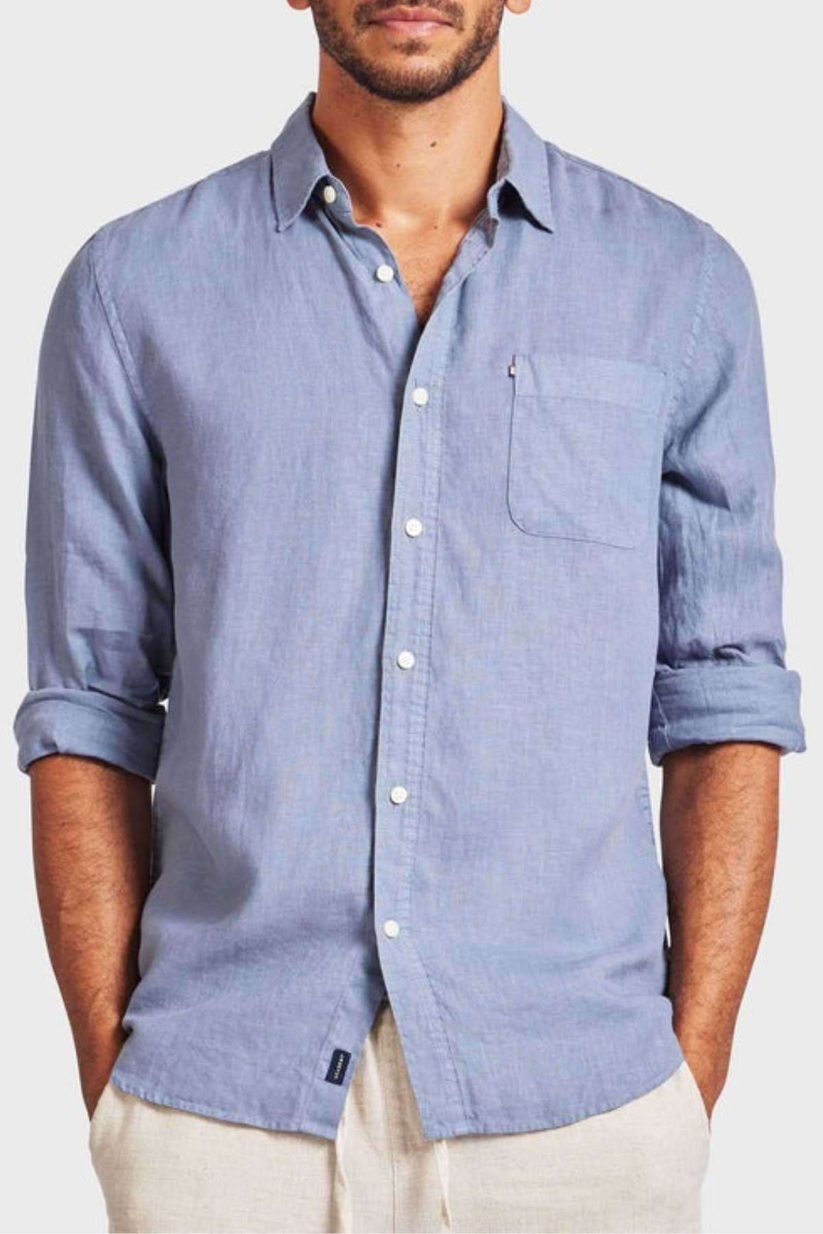 A person is wearing the Academy Brand Hampton Linen Shirt in light blue, featuring rolled-up sleeves and a chest pocket. This lightweight staple is complemented by light-colored pants against a plain white backdrop.