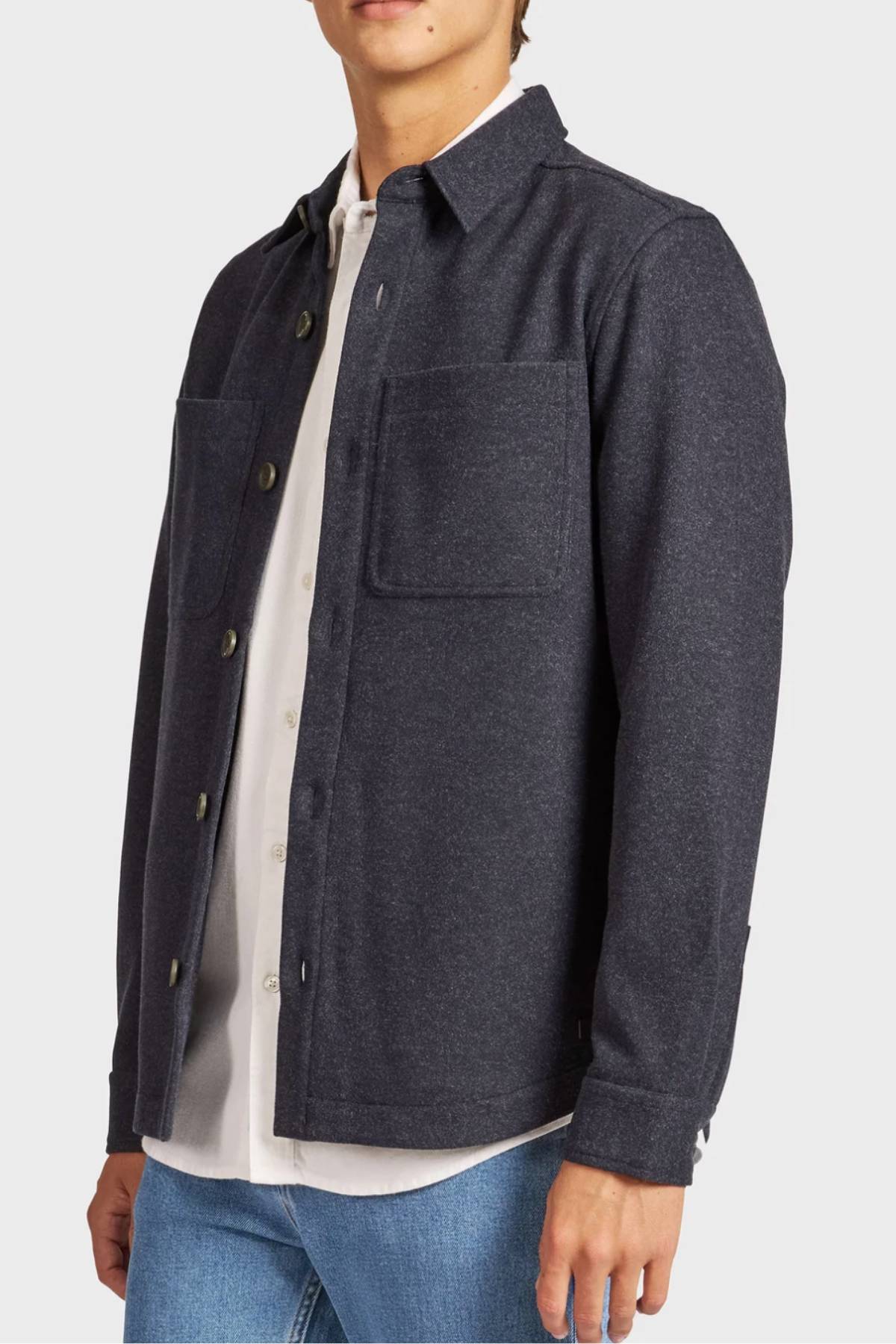 A person wears the Academy Brand Lloyd Overshirt in dark gray, featuring large breast pockets, layered over a white collared shirt and blue jeans, standing against a plain light-colored background.