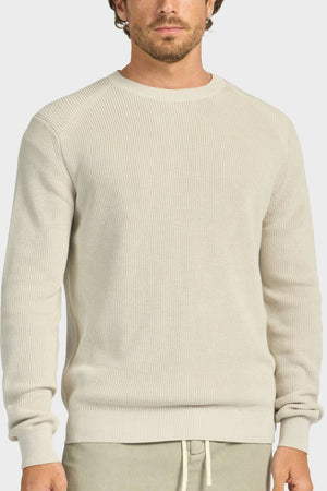 A man wearing the Academy Brand Oliver Knit Crew, a light beige ribbed cotton sweater with long sleeves and a crew neckline, paired with light pants, stands against a plain background.