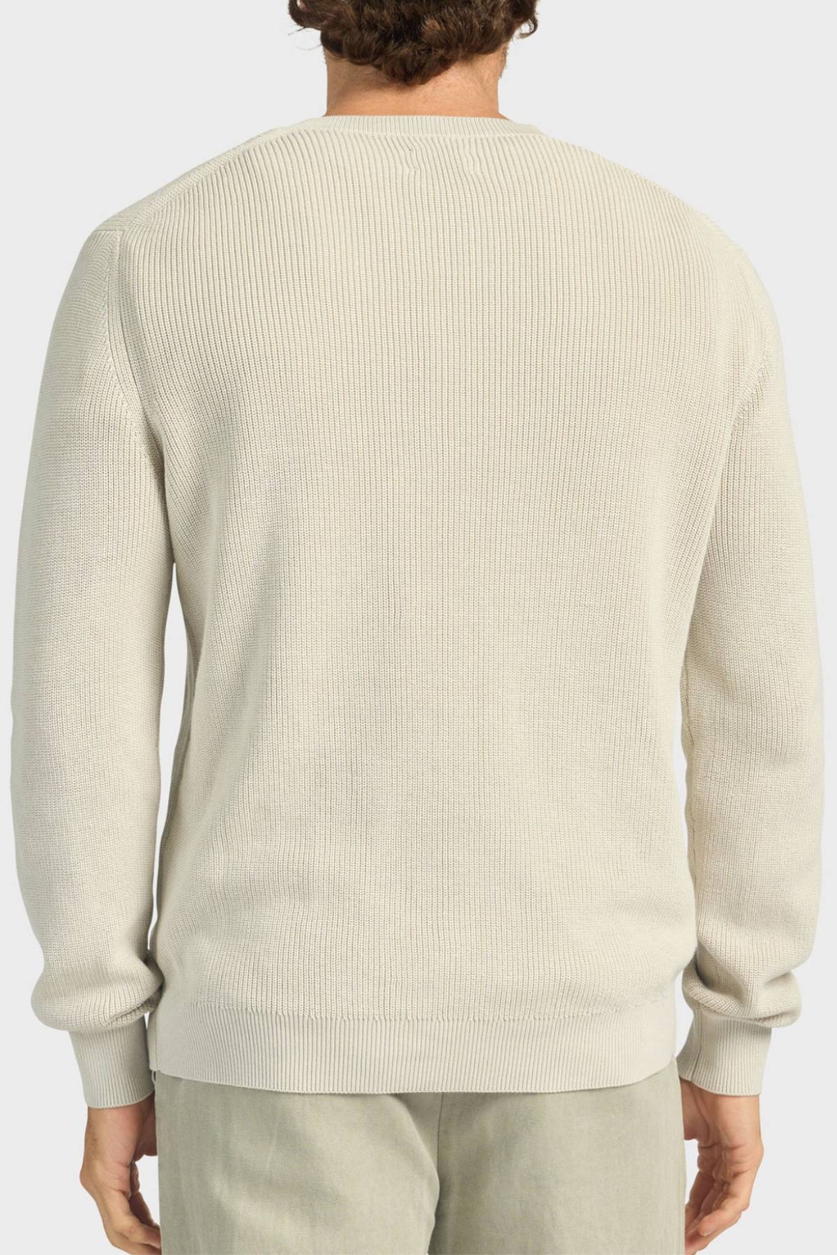 A person is seen from behind wearing the Academy Brand Oliver Knit Crew, a light beige, relaxed-fit sweater made from soft cotton yarn with a vintage wash and ribbed crew neckline. They pair it with light-colored pants against a plain white background.