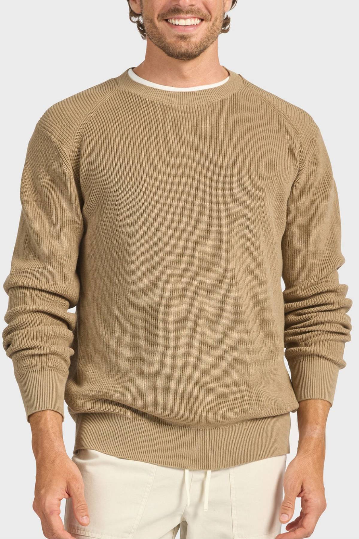 A smiling man wears the Academy Brand Oliver Knit Crew, a light brown cotton sweater, over a white shirt and light pants while standing against a plain background.