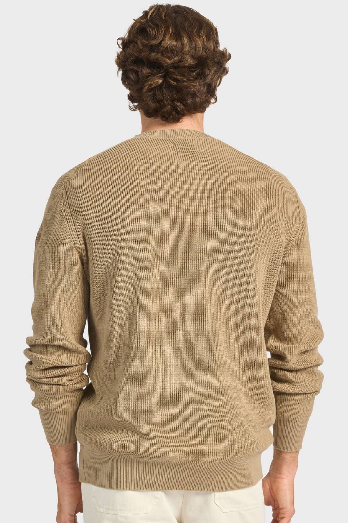 A man with wavy brown hair is shown from the back, wearing the Academy Brand Oliver Knit Crew in ribbed premium cotton yarn and white pants against a plain light background.