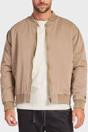 A man stands against a light background wearing the Academy Brand Standard Issue Bomber, a retro military-inspired 100% cotton twill jacket with ribbed cuffs and collar, unzipped over a white t-shirt and dark pants.