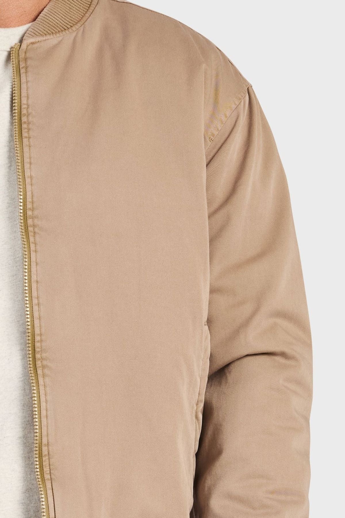 A close-up of someone in the Academy Brand Standard Issue Bomber, a light brown retro military-inspired jacket with zipper and ribbed collar over a white shirt, shown from the upper left against a plain light gray background.