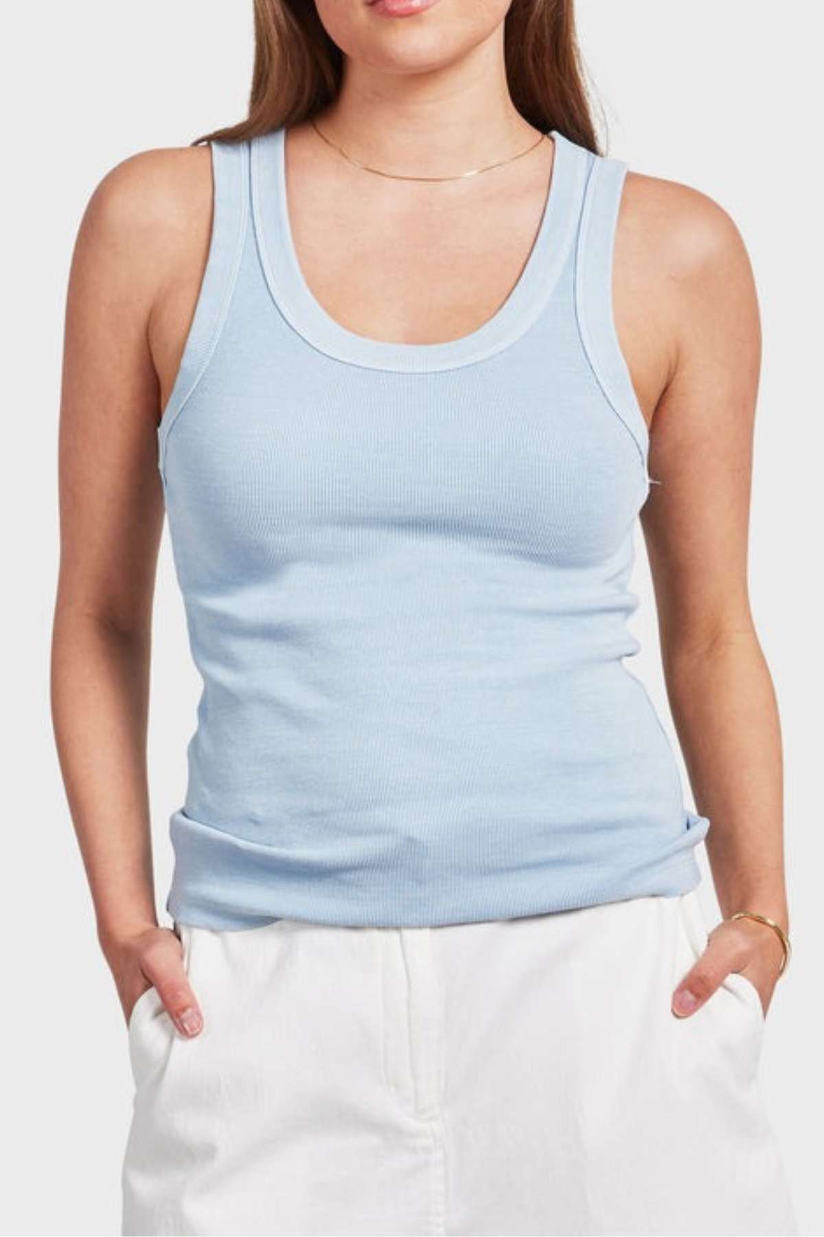 A person dressed in an Academy Women Essential Rib Tank in light blue paired with white pants, with hands casually tucked into the pockets, stands against a plain white background.