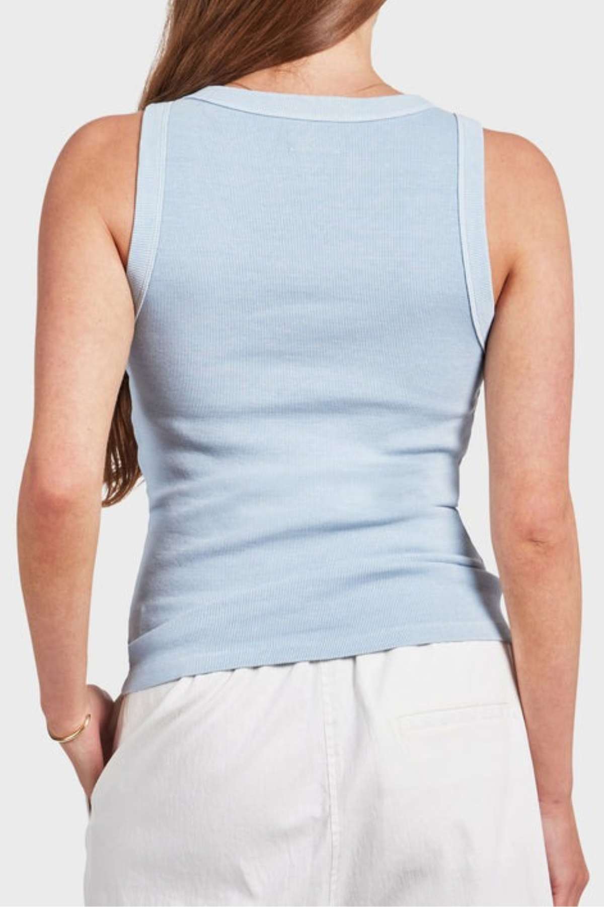 A person with long hair is wearing the comfortable Academy Women Essential Rib Tank in light blue and a pair of white pants, standing with their back to the camera. They are holding a small bag or object in their right hand.