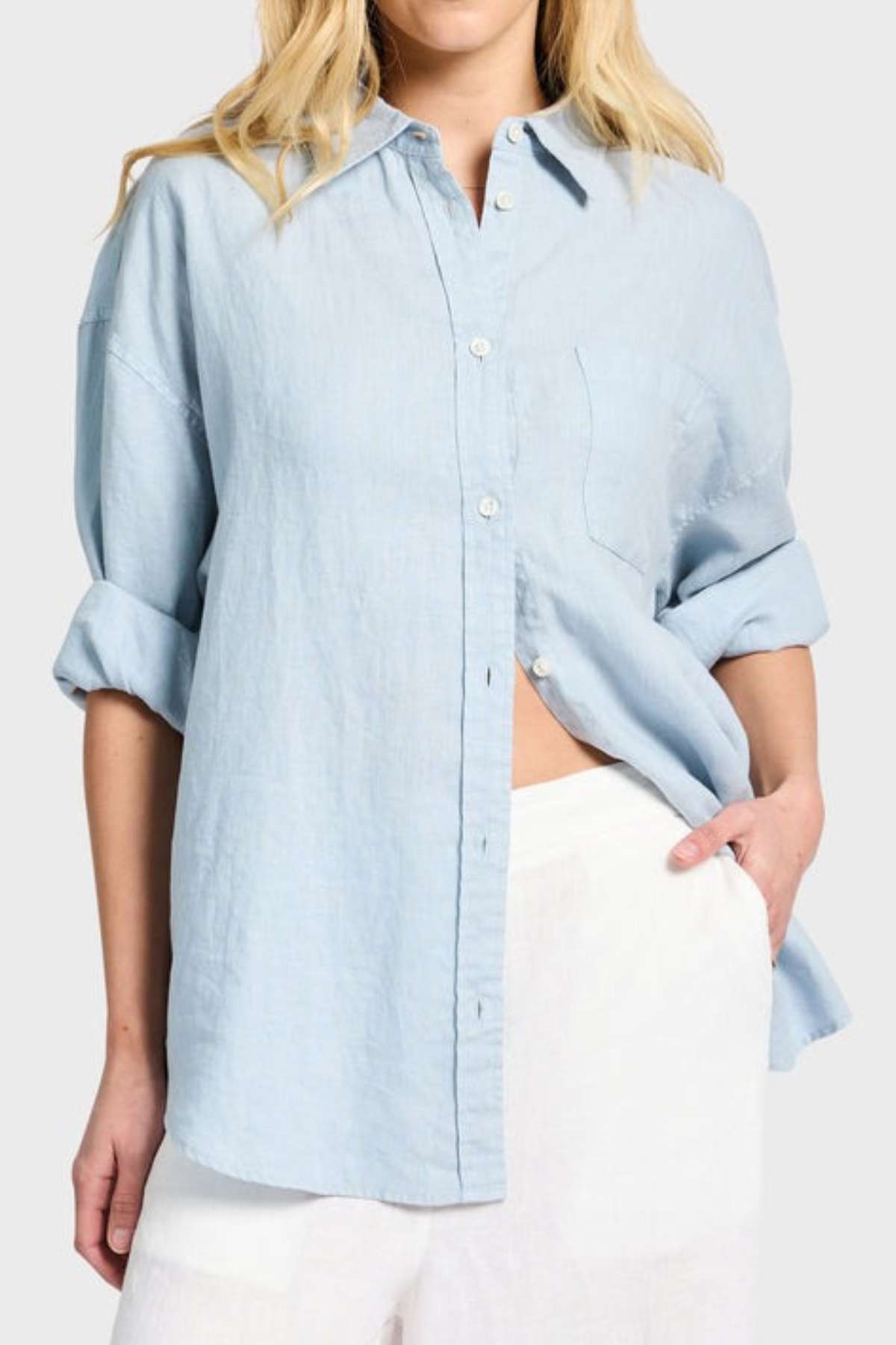 A person wearing a light blue Academy Women Hampton Linen Shirt, complete with rolled-up sleeves and mother-of-pearl buttons, pairs it effortlessly with white pants. With one hand resting in their pocket against a neutral background, they exude a relaxed yet polished vibe.