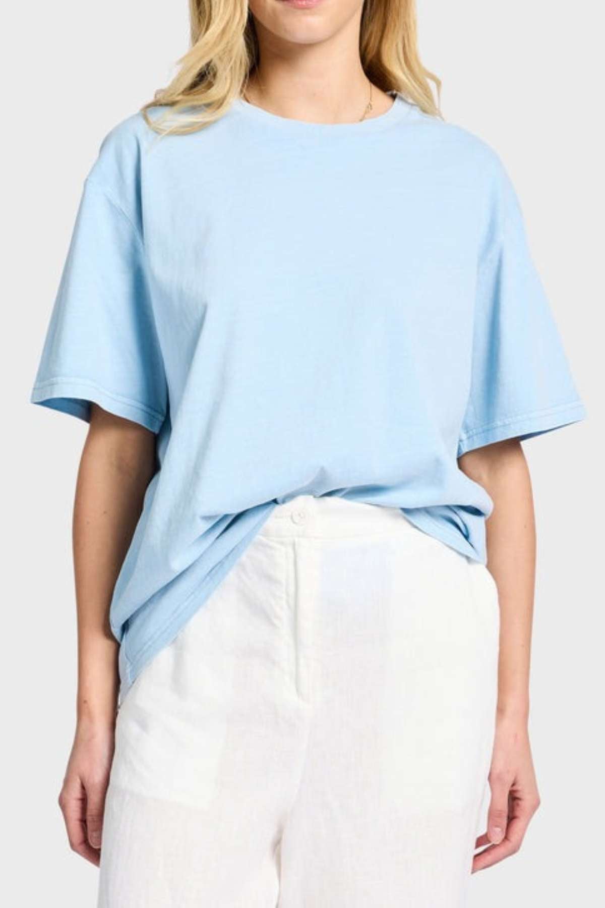 A person is wearing a light blue Academy Women Jimmy Boyfriend Tee, made from premium sanded jersey, tucked into high-waisted white pants. They stand against a plain gray background with long blonde hair cascading down, their hands resting at their sides. The outfit exudes a casual and relaxed vibe.