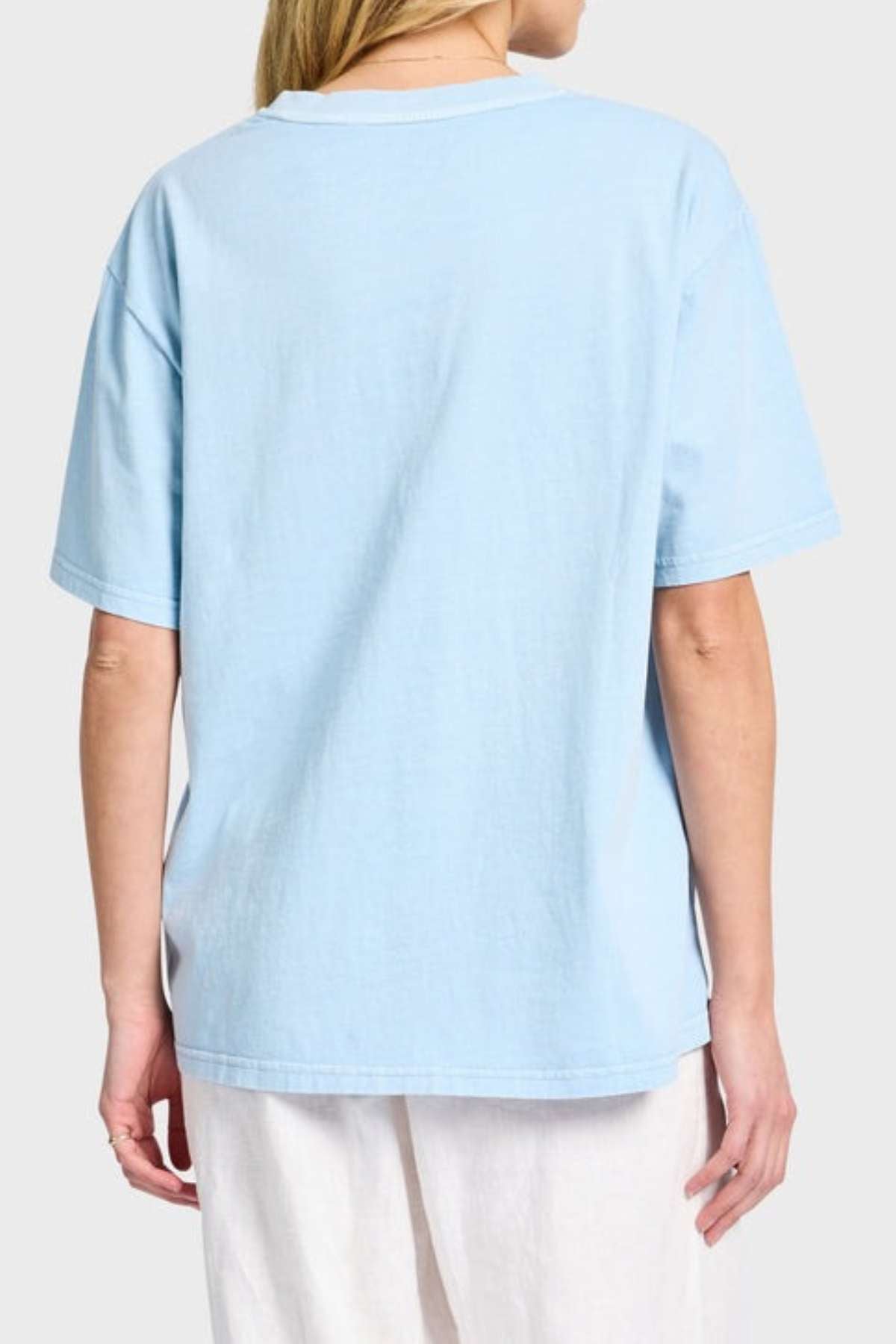A person with light hair is wearing an oversized, light blue Academy Women Jimmy Boyfriend Tee and white pants, standing with their back to the camera against a plain gray background.