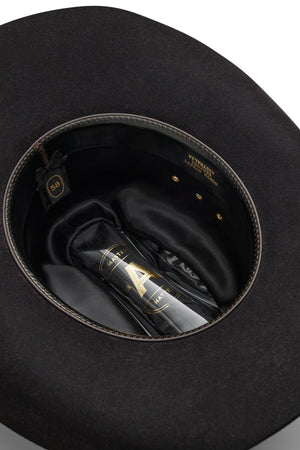 An Akubra Outback Club hat in black, size 58, sits upside down to display its inner lining and a clear bottle with gold and black labeling nestled in the crown—a tribute to Lee Kernaghan and Australian country music.