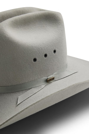 A close-up of a light grey Akubra Outback Club hat features a matching ribbon band, three ventilation holes, and a small metal AKUBRA tag on the ribbon. Inspired by Australian country music legends. White background.