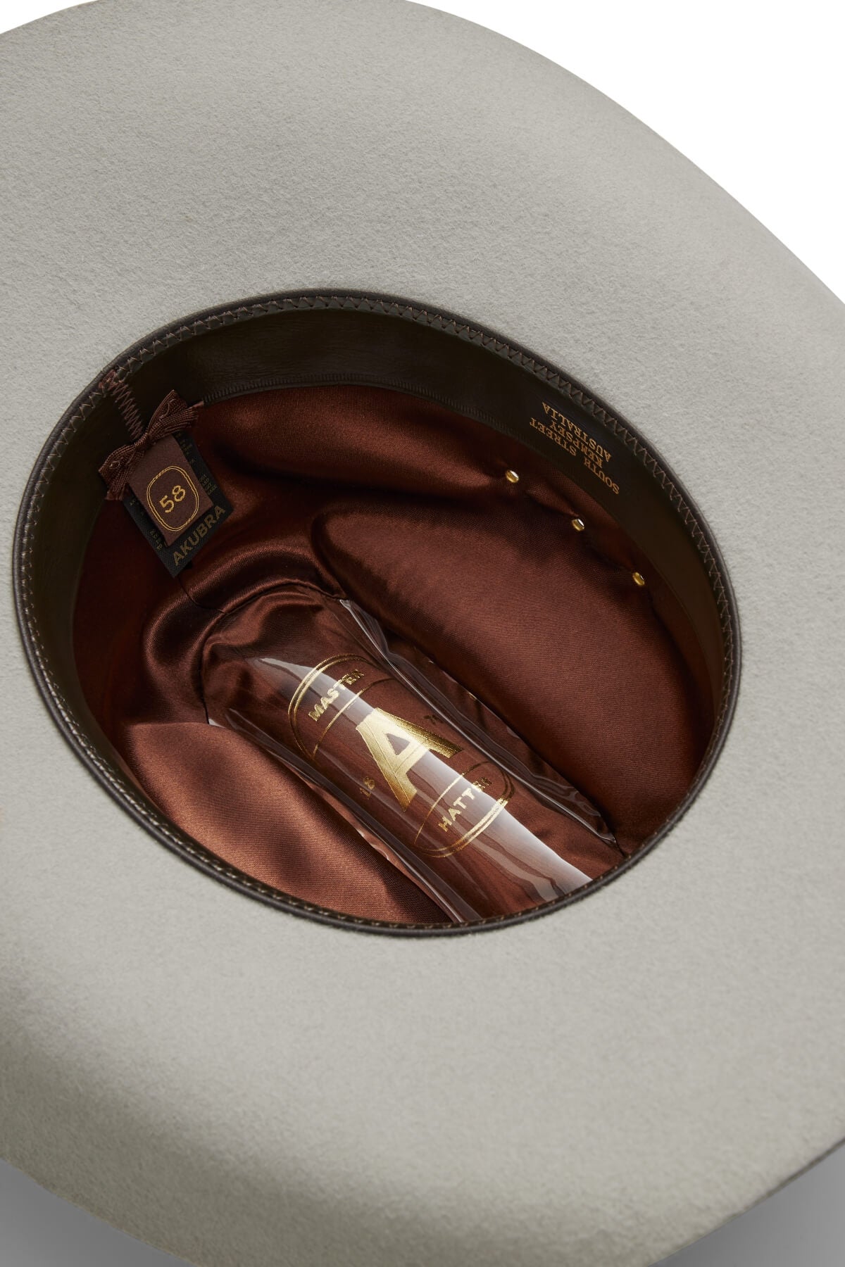 A top-down view of the inside of a beige Akubra Outback Club hat with a brown satin lining and a size label reading "59." Inside, there's a clear bottle labeled "AURA" with a gold "A," capturing classic Australian country music style.