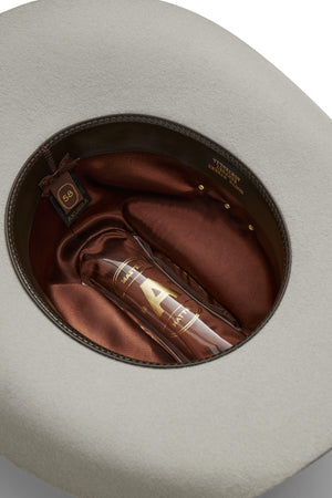 A top-down view of the inside of a beige Akubra Outback Club hat with a brown satin lining and a size label reading "59." Inside, there's a clear bottle labeled "AURA" with a gold "A," capturing classic Australian country music style.