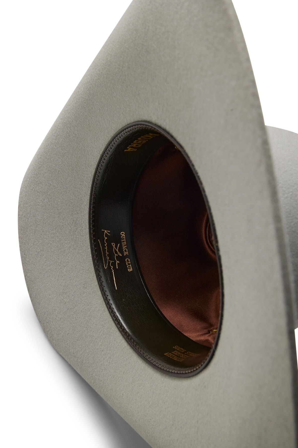 A close-up of the inside of a light gray Akubra Outback Club hat, featuring a brown satin lining and a black leather sweatband with gold writing and a Lee Kernaghan signature, celebrating Australian country music style.