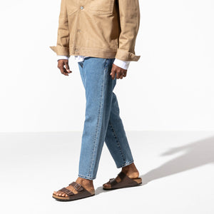 A person in a tan jacket, light blue jeans, and brown Birkenstock Arizona Birko-Flor Regular sandals walks against a plain white background. Their face and upper body are not visible in the image.