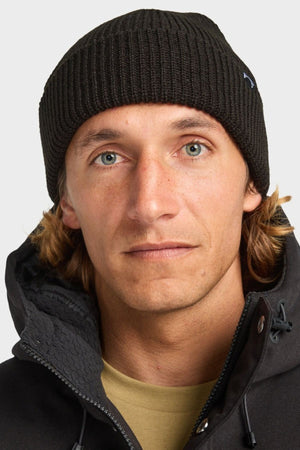 A man with light eyes and shoulder-length blond hair wears a black Billabong Arcade Beanie in acrylic gauge rib knit and a black jacket, looking directly at the camera with a neutral expression against a plain background.